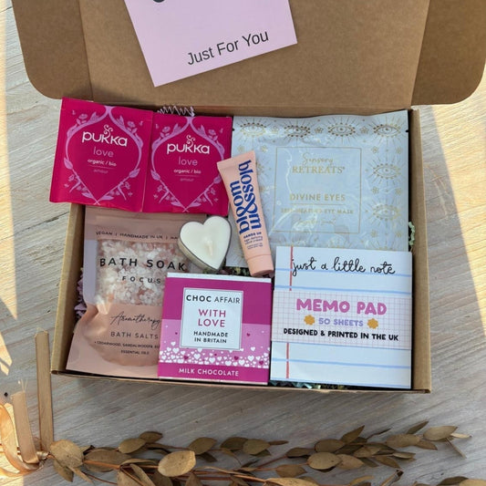 One Box of Happiness gift box with pink Pukka tea, lavender eye mask, heart candle, bath soak, chocolate bar, and memo pad.